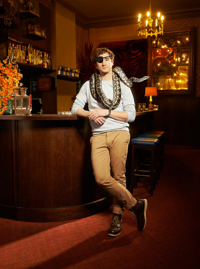 Adult male in light blue sweater and khakis poses next to a bar with a snake around his shoulders in an indoor environment with warm lighting.