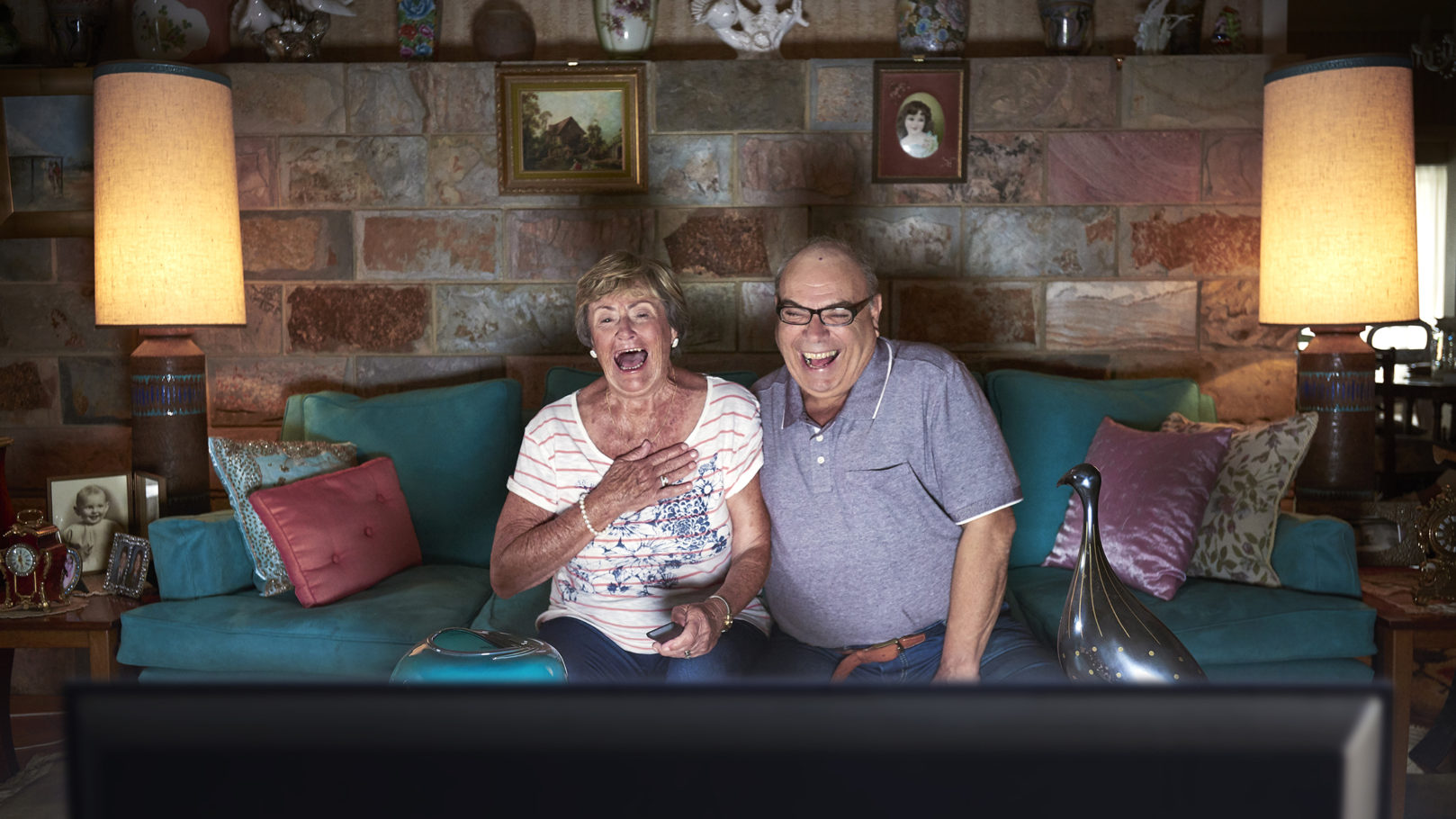 Elderly couple laughing and watching TV in a cozy living room with stone walls, lamps, decorative objects, and a turquoise couch.