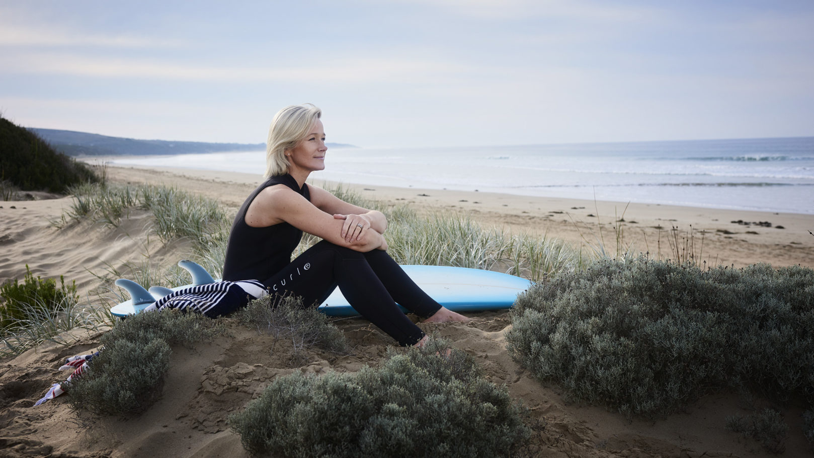 Adult woman in wetsuit sits on sandy beach with surfboard looking out at ocean under cloudy sky, enjoying the scenic view.