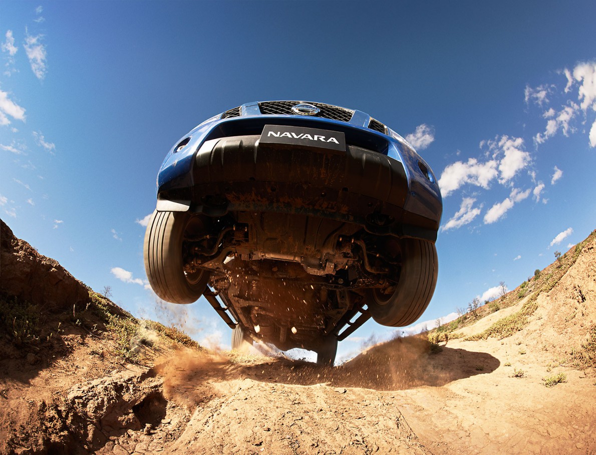 Blue Nissan Navara mid-air on a dirt road under a blue sky, captured from a low angle emphasizing the vehicle's off-road capabilities.