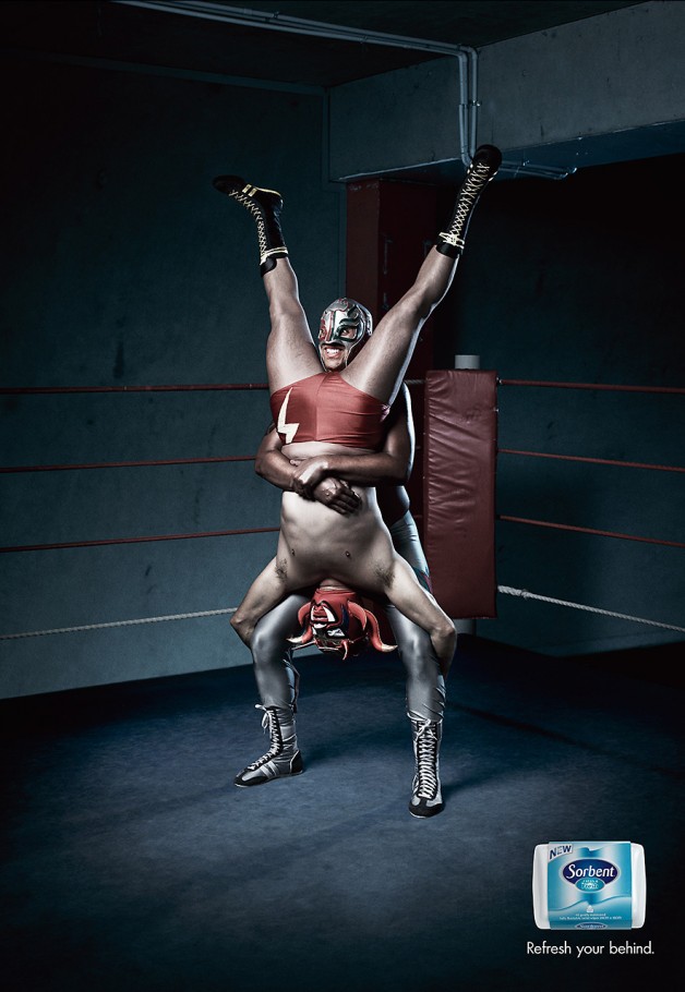 Two masked wrestlers perform an acrobatic wrestling move on a blue mat in a boxing ring, an advertisement for Sorbent tissue.