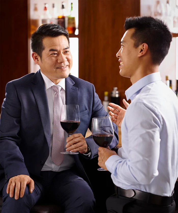 Two Asian men in a bar holding glasses of red wine while talking and smiling together