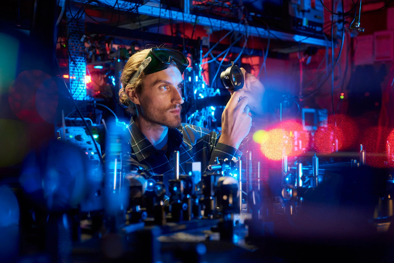 A man in safety goggles examining a piece of optical equipment in a laboratory lit with blue and red light.