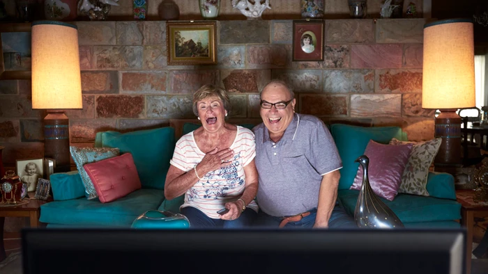 An elderly couple laughing while seated on a turquoise sofa in a living room, photographed by Stuart Crossett.