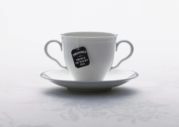White tea cup with two handles and a saucer featuring a Twinings Prince of Wales tea bag against a white background