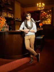 Man wearing an eyepatch and a snake wrapped around his shoulders leaning against a wooden bar in a pub.
