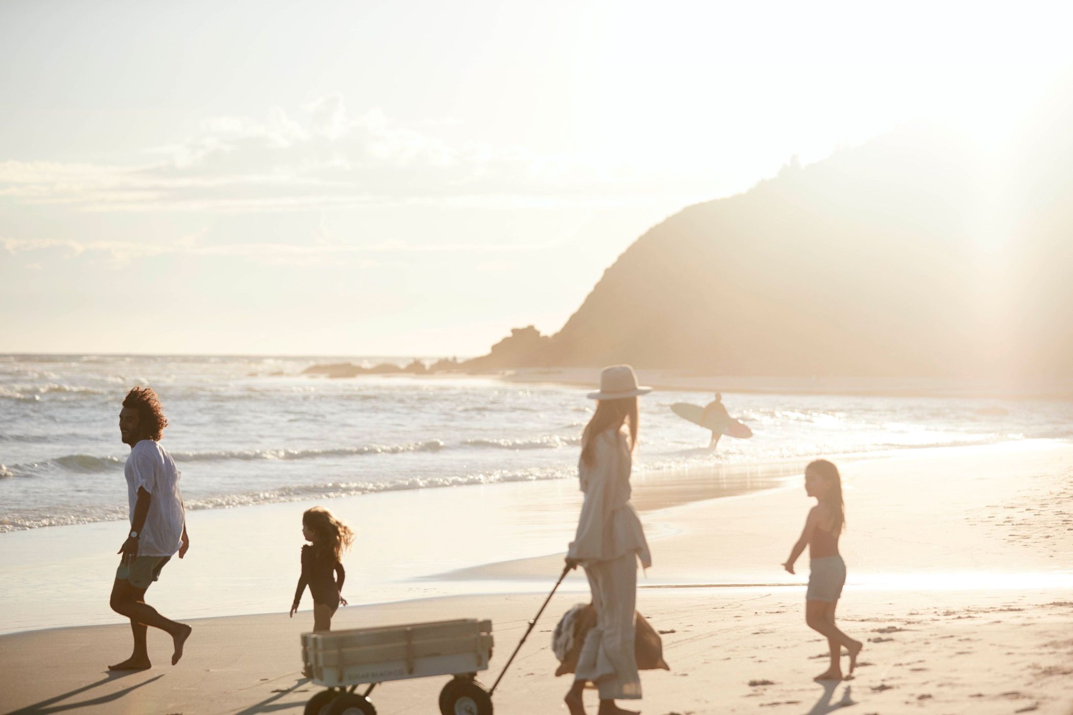 Family enjoys a sunny beach day. Mother pulls wagon with child, while father and other child walk along the shoreline near the water.