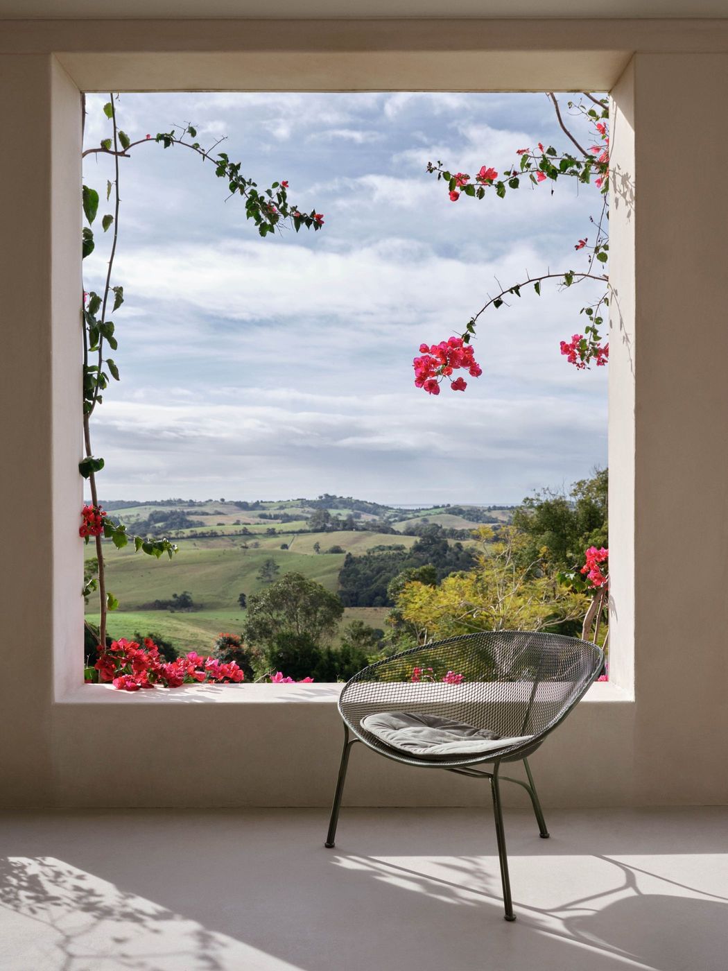 Interior view through window to a green rural landscape with chair and bougainvillea framing the scene. Interior design.