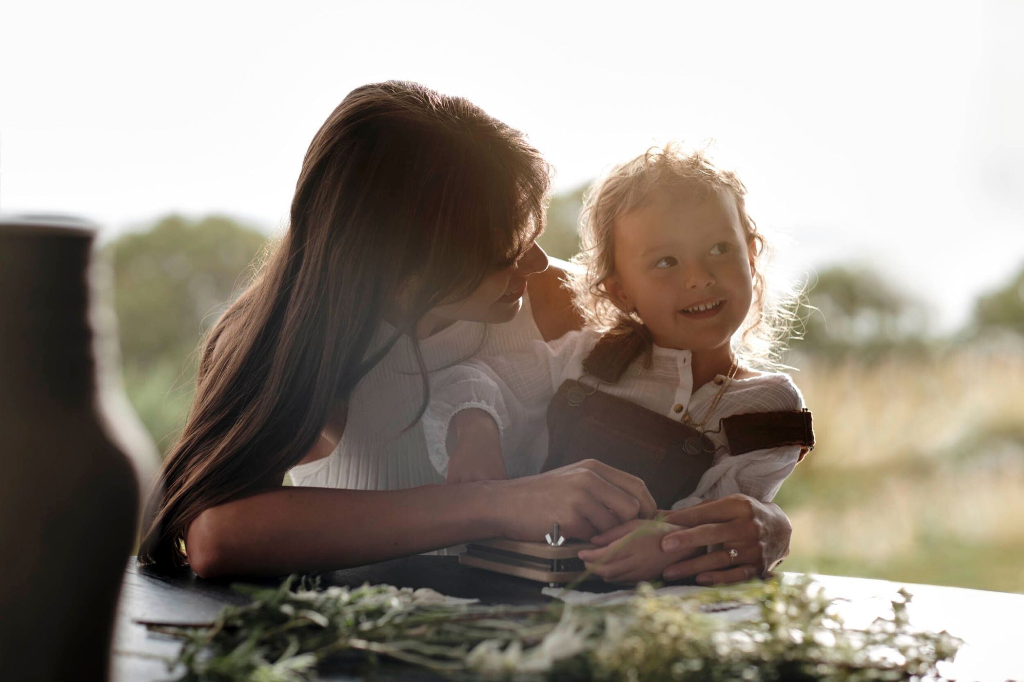 Mother and child smile outdoors at a table with herbs, backlit by natural light, showcasing a tender moment between family members.