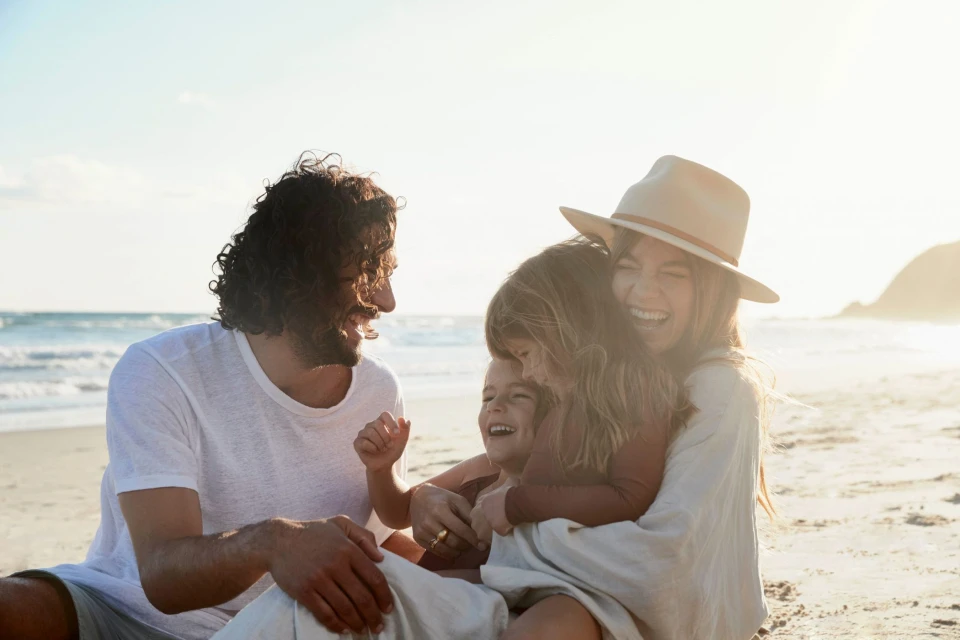 Happy family of four laughing together on a sandy beach, with a man, woman wearing a hat, and two children enjoying a sunny day.