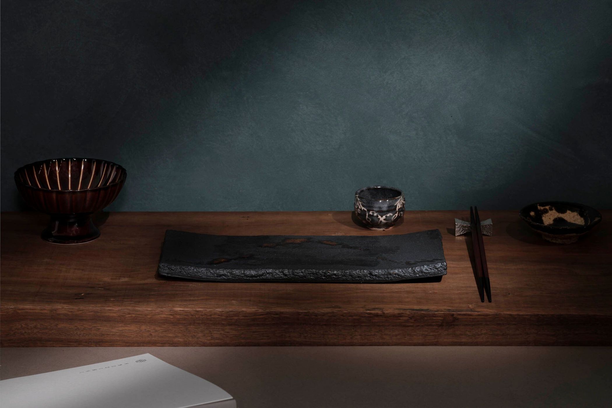 Dramatic moody food photography: Dark pottery and serving ware on a wooden surface with muted, dark teal backdrop and soft, directional lighting.