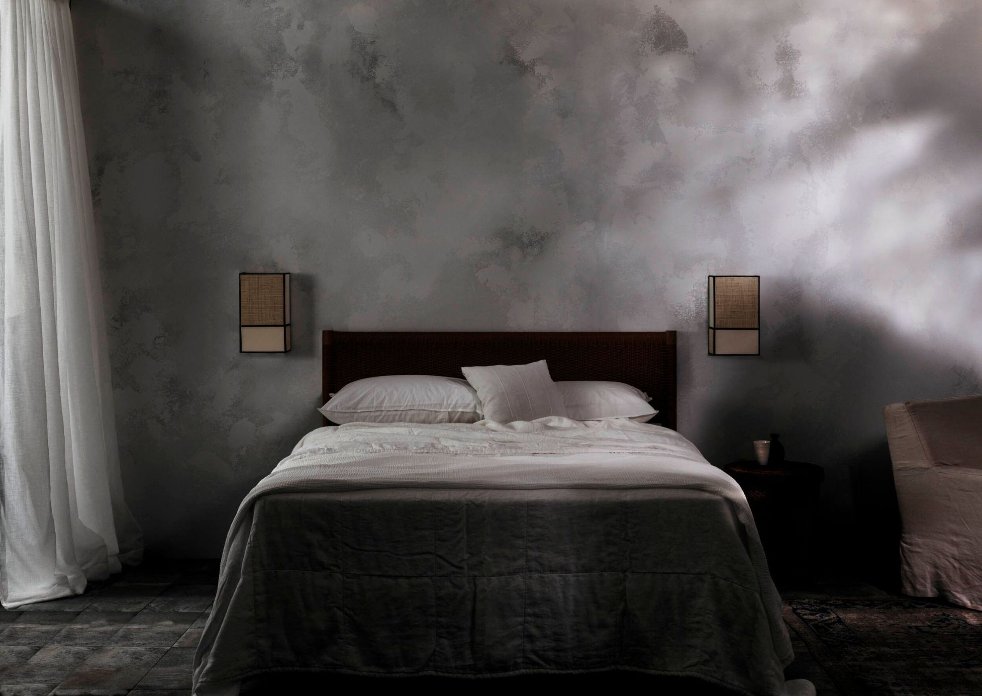 Bedroom interior featuring a linen-covered bed with pillows, dark wooden headboard, two sconces, and grey mottled wall with natural light.
