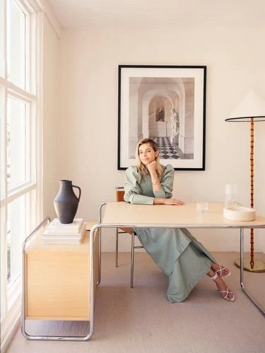 Woman in a sage green dress sits at a desk with a vase, books, and framed art in an interior space with neutral colors and natural light.