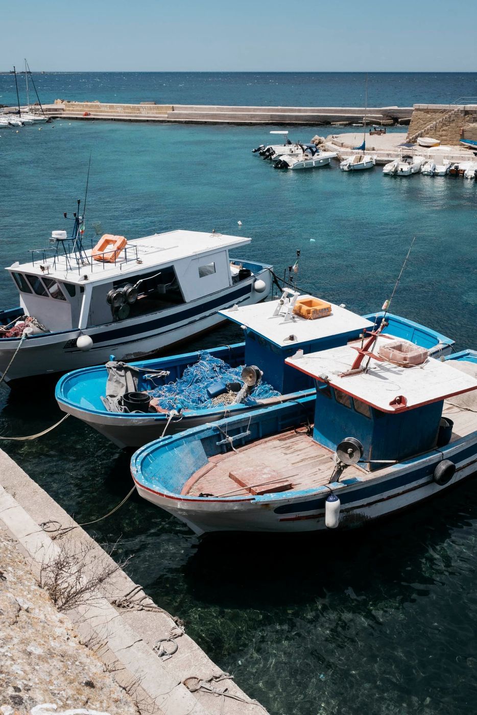 Tranquil harbor scene featuring a trio of fishing boats docked alongside a stone pier, with clear turquoise water and a distant breakwater.
