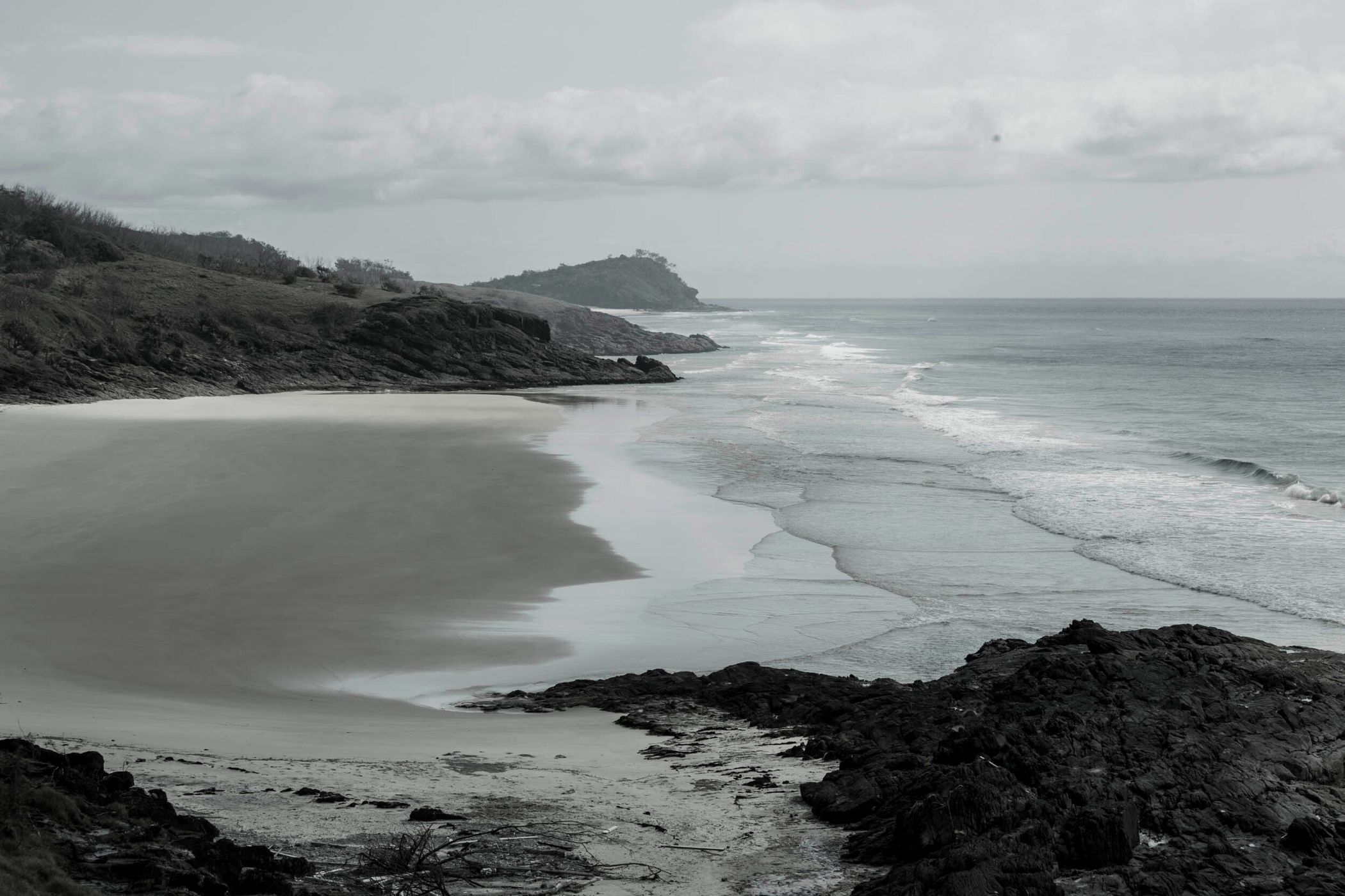 Monochromatic coastal scene featuring a sandy beach meeting a grey ocean under an overcast sky, with rocky outcrops framing the view.