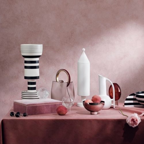 Elegant still life composition with a pale pink handbag, decorative ceramic vessels, peaches, jewelry and a velvet covered box on a pink tablecloth.