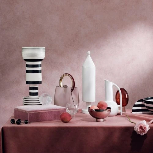 Elegant still life composition with a pale pink handbag, decorative ceramic vessels, peaches, jewelry and a velvet covered box on a pink tablecloth.