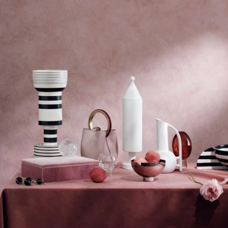 Elegant still life composition with a pale pink handbag, decorative ceramic vessels, peaches, jewelry and a velvet covered box on a pink tablecloth.