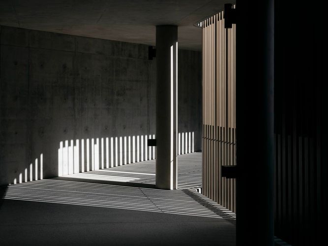 Architectural photography of a modern interior with light shafts illuminating concrete walls and columns, creating shadows and geometric patterns.
