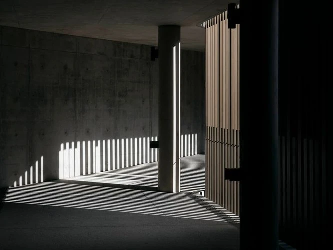 Architectural photography of a modern interior with light shafts illuminating concrete walls and columns, creating shadows and geometric patterns.