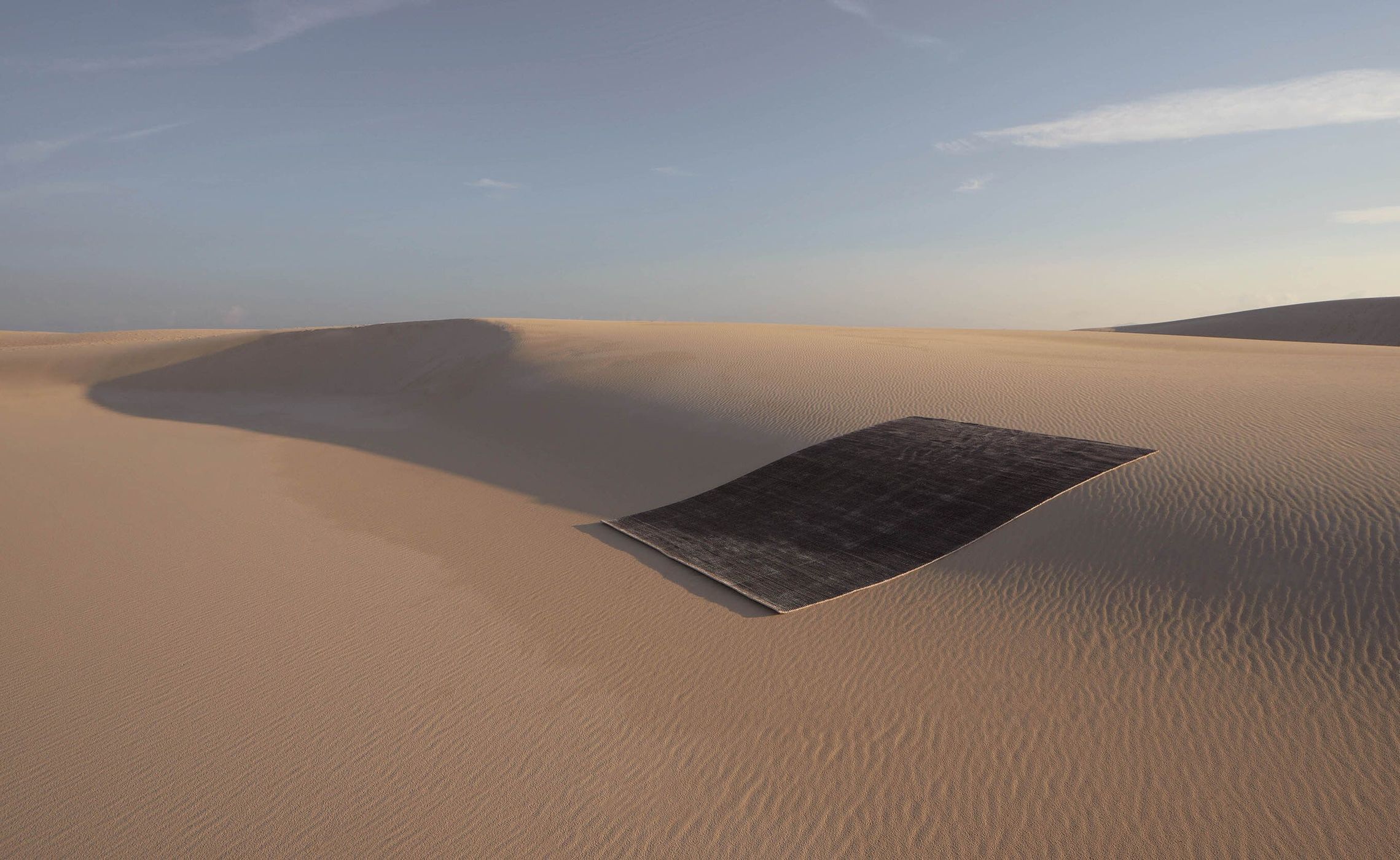 Abstract photograph of a black rug placed on a large sand dune in a desert landscape with a light blue sky overhead.