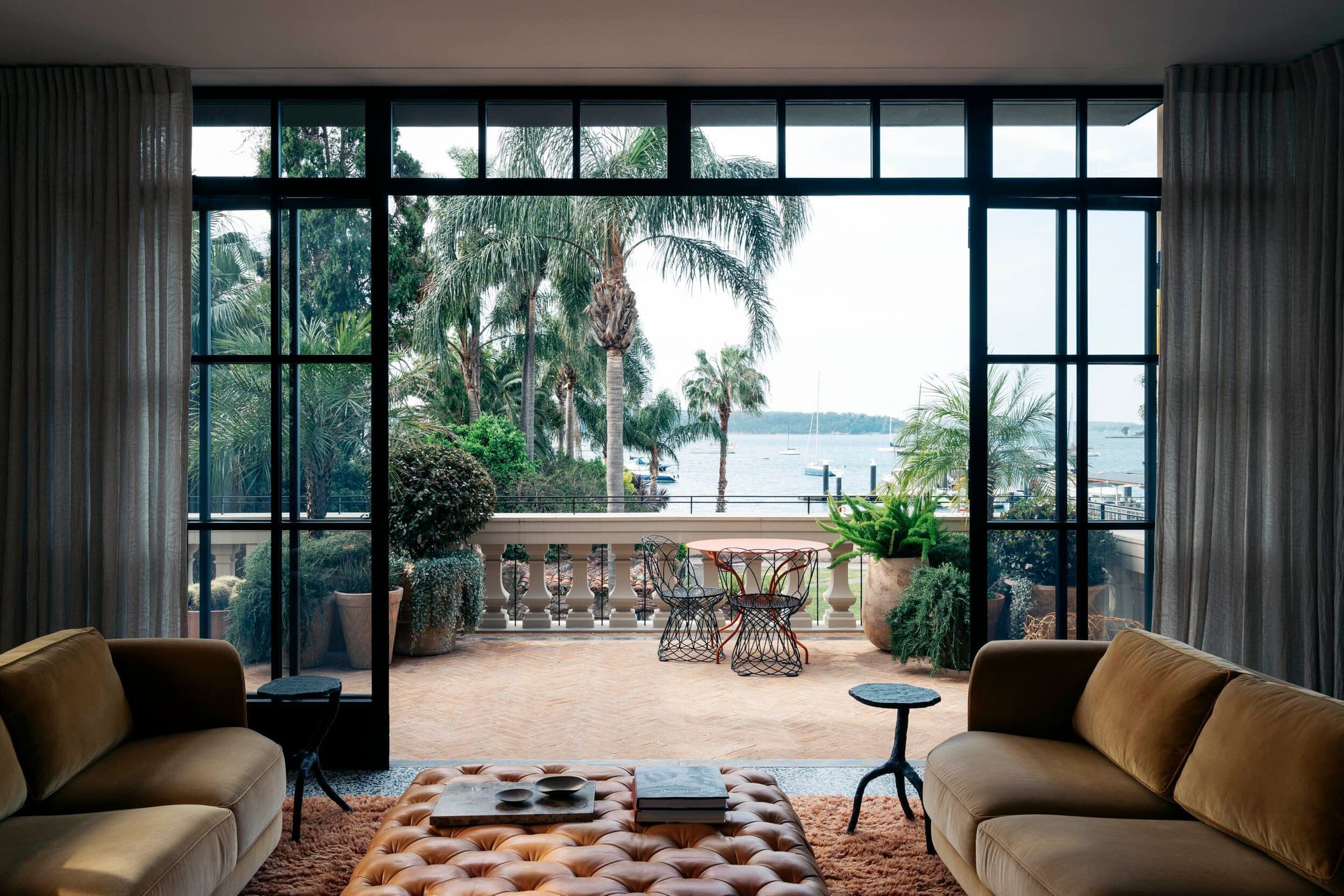 Living room with a view of a patio, waterfront, and palm trees through a large black-framed window, furnished with sofas and an ottoman.