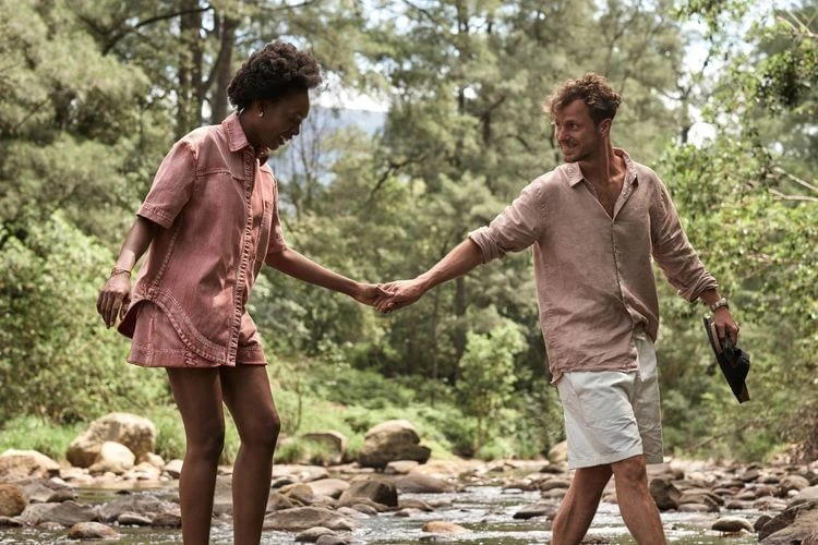 A couple walks through a rocky riverbed surrounded by a forest. The woman wears a pink short set, and the man wears white shorts and a beige shirt.