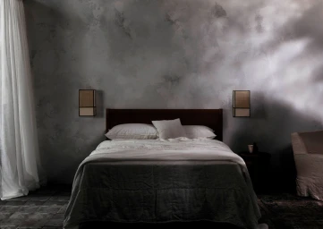 Minimalist bedroom interior with a dark wooden bed, white linens, and textured grey walls photographed by Felix Forest.