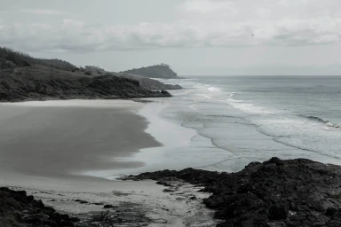 Monochrome landscape photograph of a rugged coastline with a sandy beach and gentle ocean waves rolling in under an overcast sky.