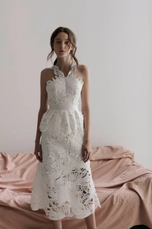 Young female model wearing a white laser-cut patterned dress standing in front of a draped pink fabric background.