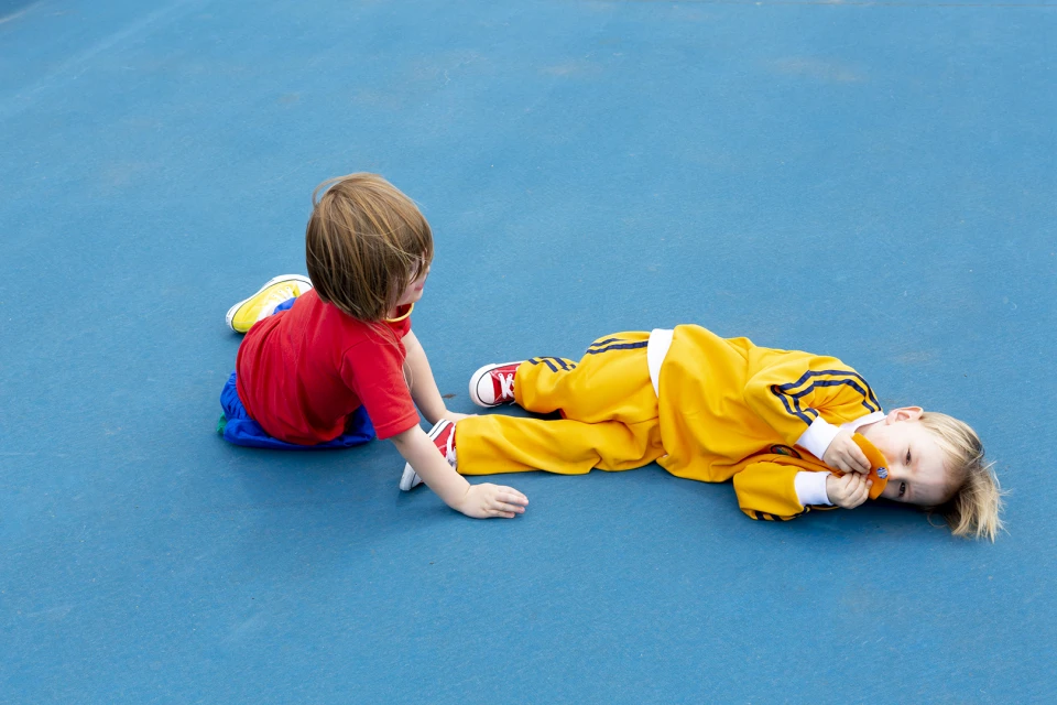Two young children, one in yellow tracksuit lying on the ground and other seated in red shirt on blue surface outdoors.