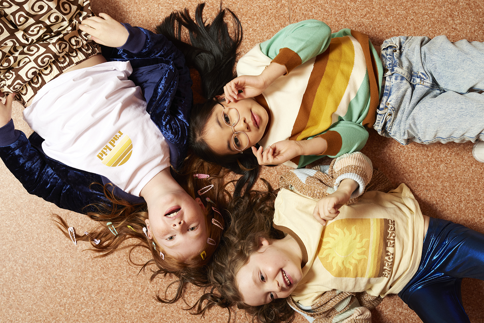 Three smiling young girls lie in a circle on the floor in a playful pose wearing vintage-inspired outfits with soft, diffused lighting.