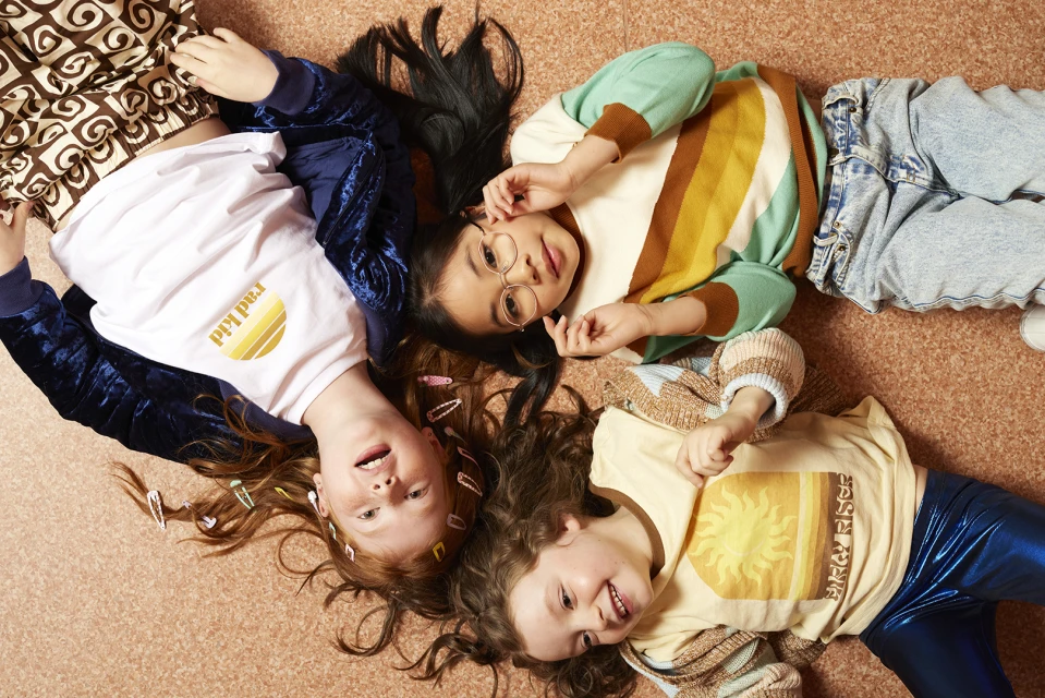 Three smiling young girls lie in a circle on the floor in a playful pose wearing vintage-inspired outfits with soft, diffused lighting.