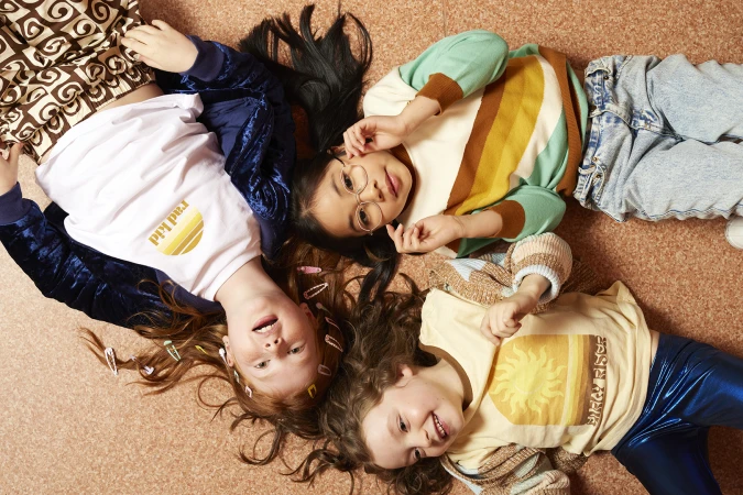 Three smiling young girls lie in a circle on the floor in a playful pose wearing vintage-inspired outfits with soft, diffused lighting.