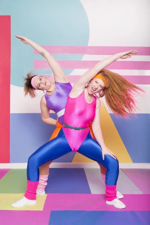 Two women in brightly colored 1980s-style exercise outfits perform a side stretch in a photo studio with a geometric backdrop.