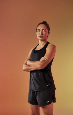 Portrait of a woman wearing black athletic tank top and shorts with arms crossed against gradient background.