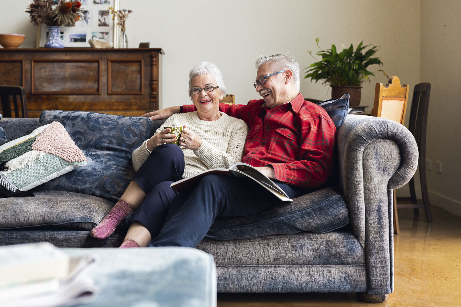 Elderly couple relaxing indoors on a couch with a book and tea, smiling and laughing in a warmly lit, comfortable home setting.