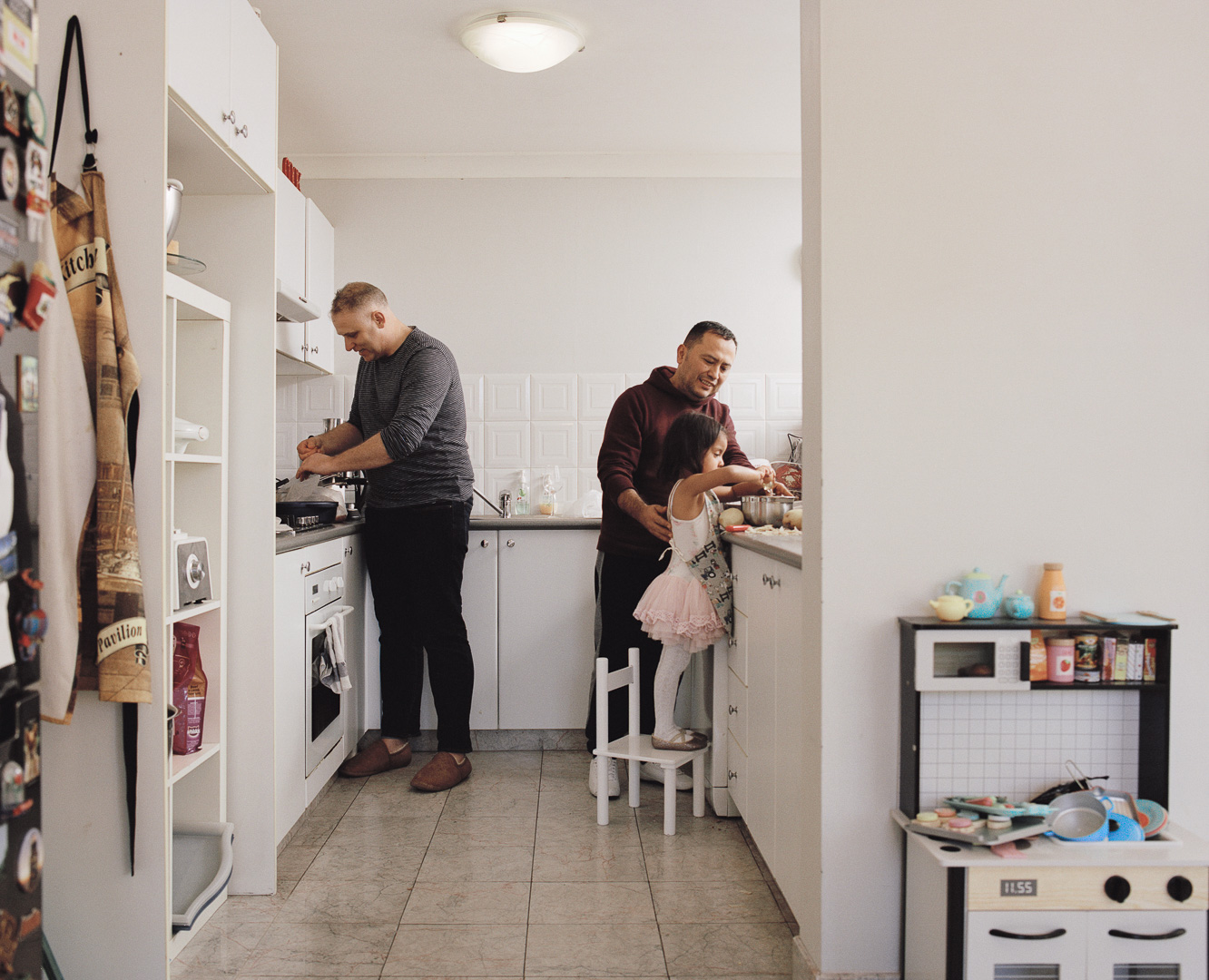 Two men and a young child prepare food in a bright, white kitchen. Kitchen appliances and a toy kitchen set are visible.