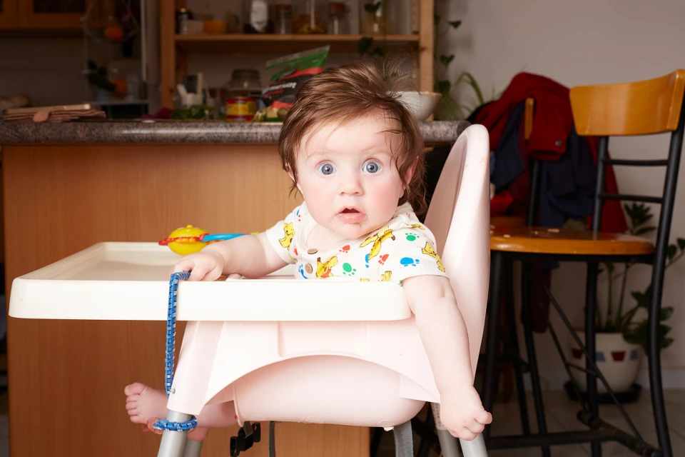 Baby sits in a high chair with a surprised facial expression, wearing a colorful shirt with a kitchen and plants in the background.