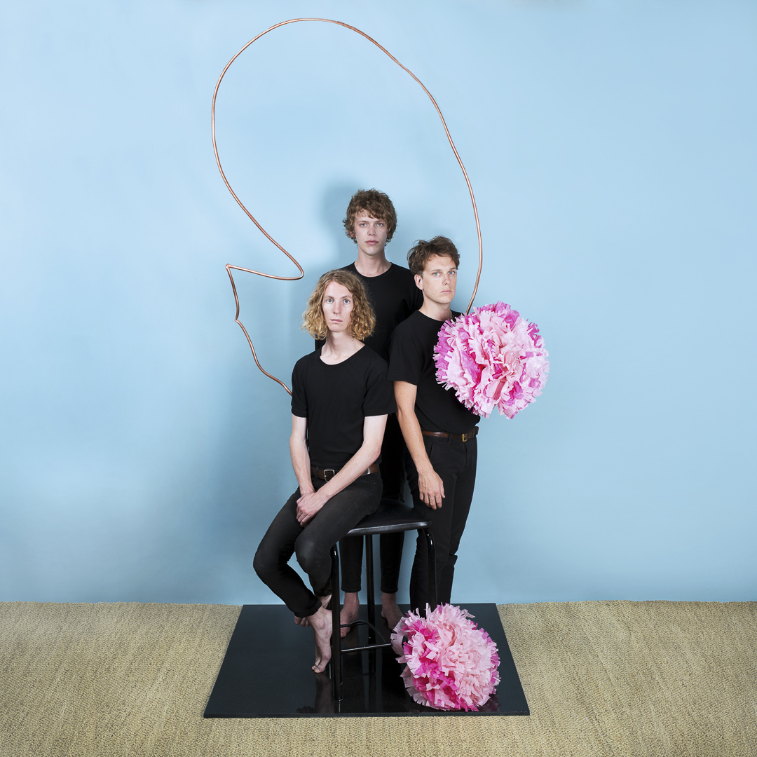 Three young adults in black attire pose indoors with paper flower props and a copper wire sculpture against a light blue background.