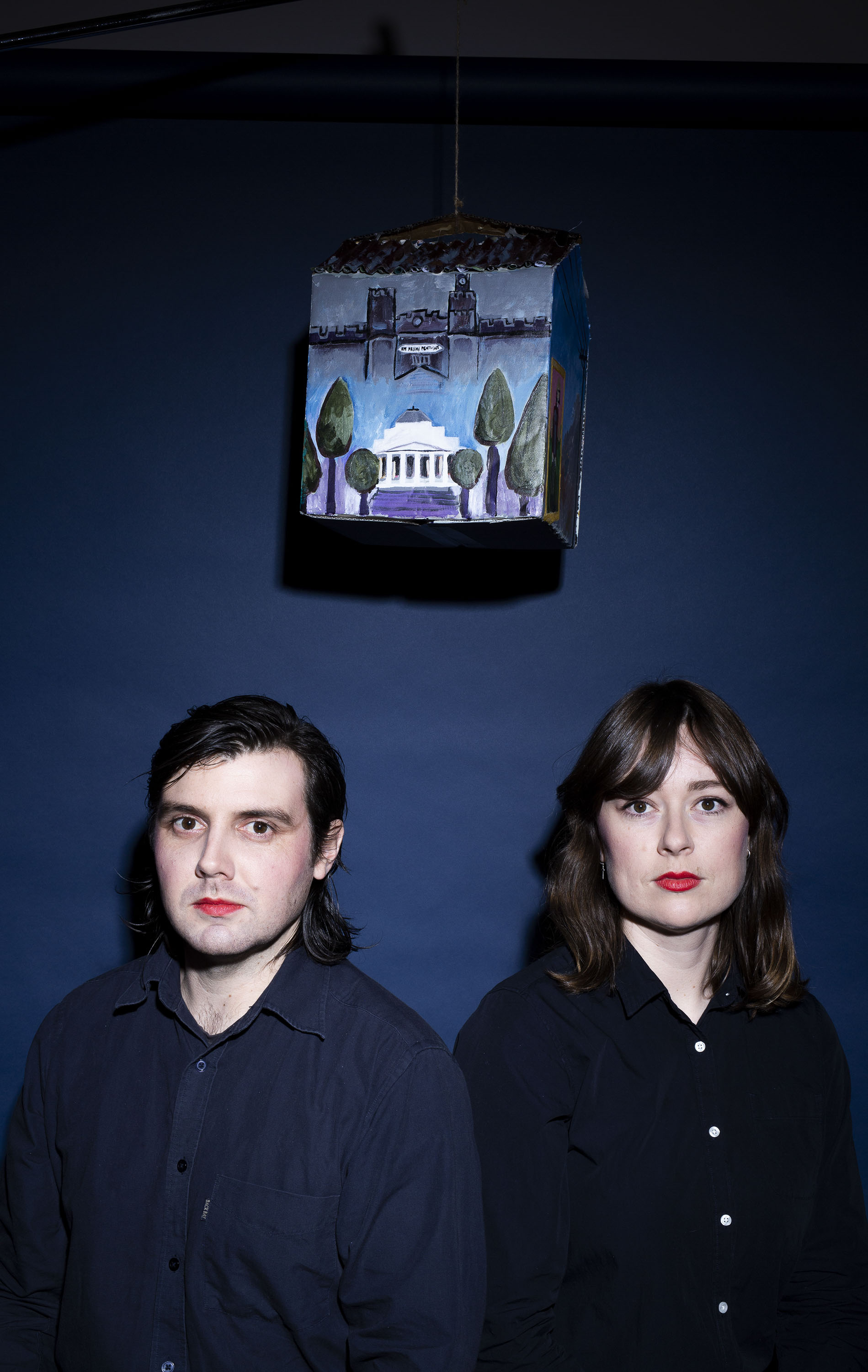 Portrait of a man and a woman with red lipstick and dark clothes beneath a hanging, painted birdhouse against a blue backdrop.