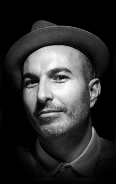 Black and white portrait of a man wearing a fedora hat looking at the camera in a studio with dramatic lighting.