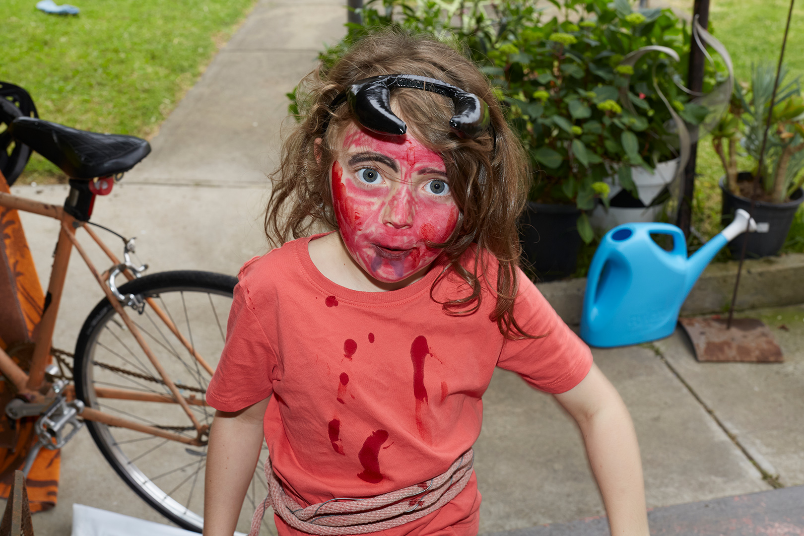Child with red face paint, goggles on head, stands outside near bicycle and greenery, wearing a coral t-shirt with rope belt.