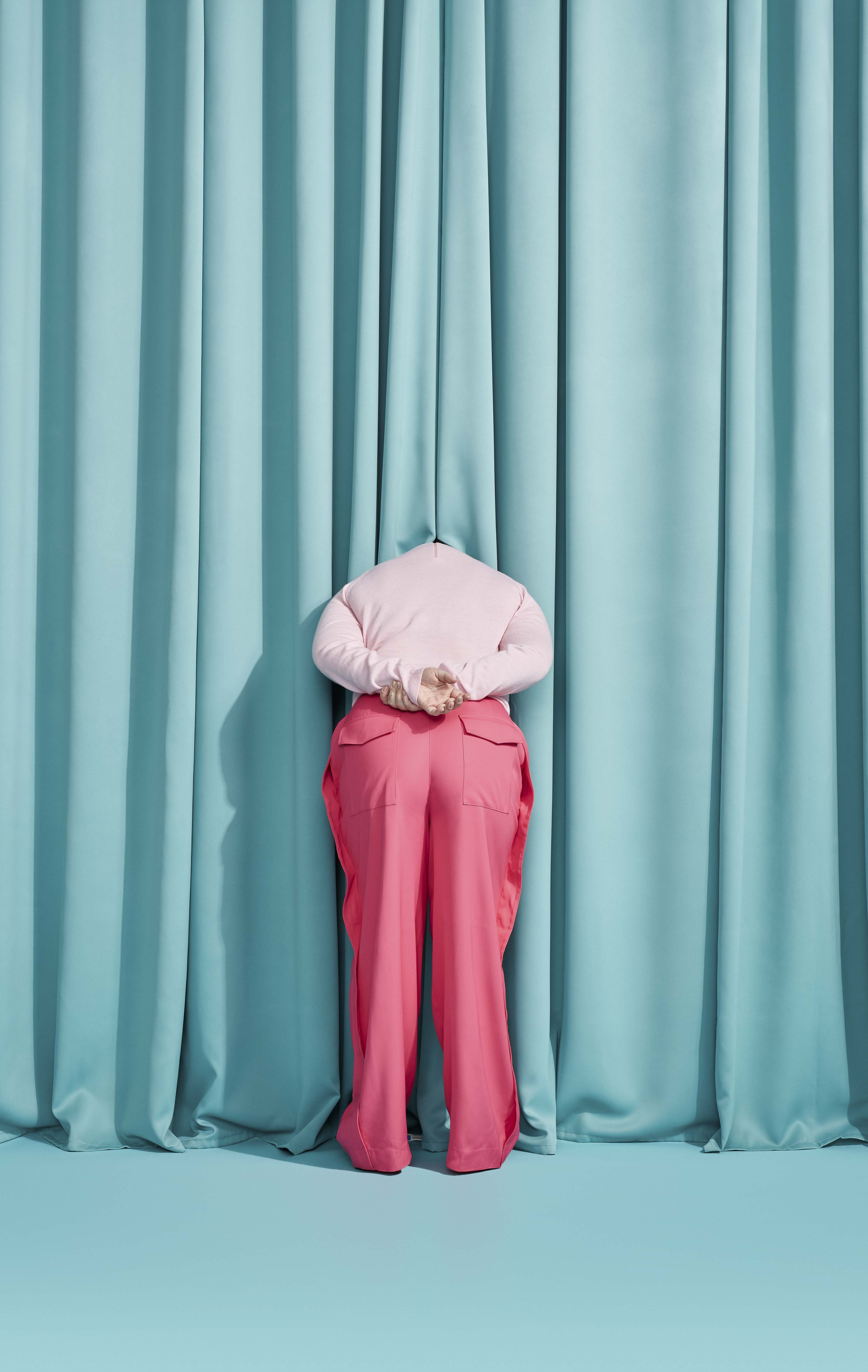 Person facing a light blue curtain wearing pink pants and a light pink shirt with their hands folded behind their back.