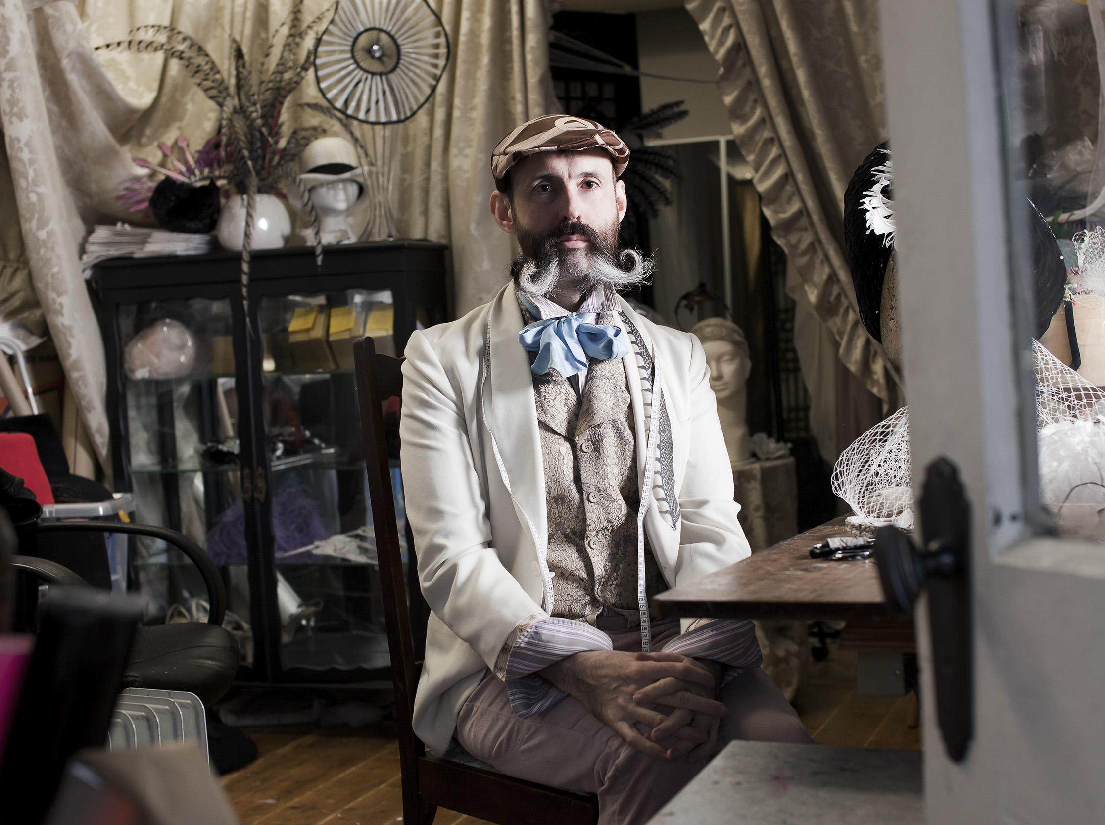 Adult male seated indoors wearing a hat, white jacket, vest, and bowtie with a meticulously styled mustache in an eclectic setting.