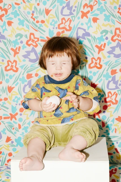 Child sits on a white block, holding a ball, wearing patterned shirt and green pants against a floral wallpaper backdrop. 