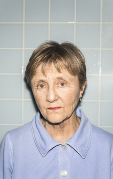 Portrait of a senior adult woman with short brown hair, wearing a blue collared shirt, posing in front of a light blue tile background.