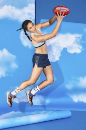 Female athlete mid-air jumping with an Australian rules football against a blue sky studio backdrop.