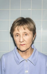 Portrait of an elderly woman with short brown hair wearing a light blue collared shirt against pale blue tiles.