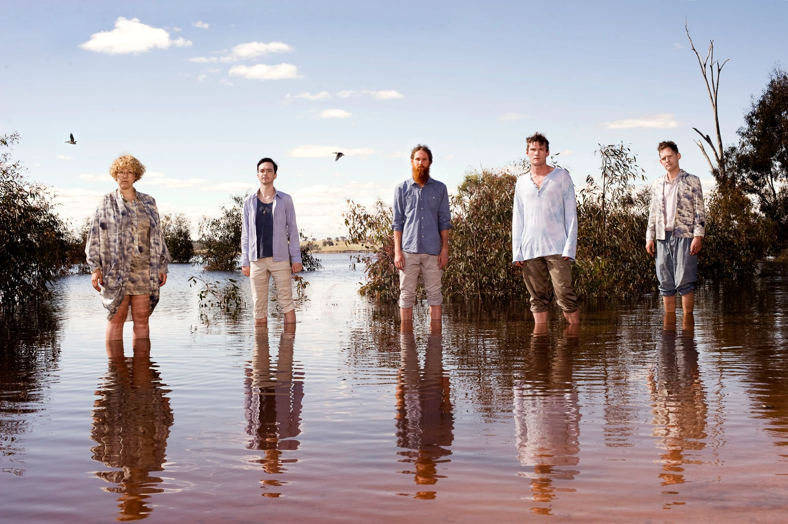 Five people standing waist-deep in shallow water of a reflective lake or flooded landscape under a blue sky.